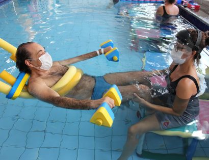 Homem com bóias em uma piscina, sendo auxiliado por uma profissional da Fisioterapia Aquática AACD