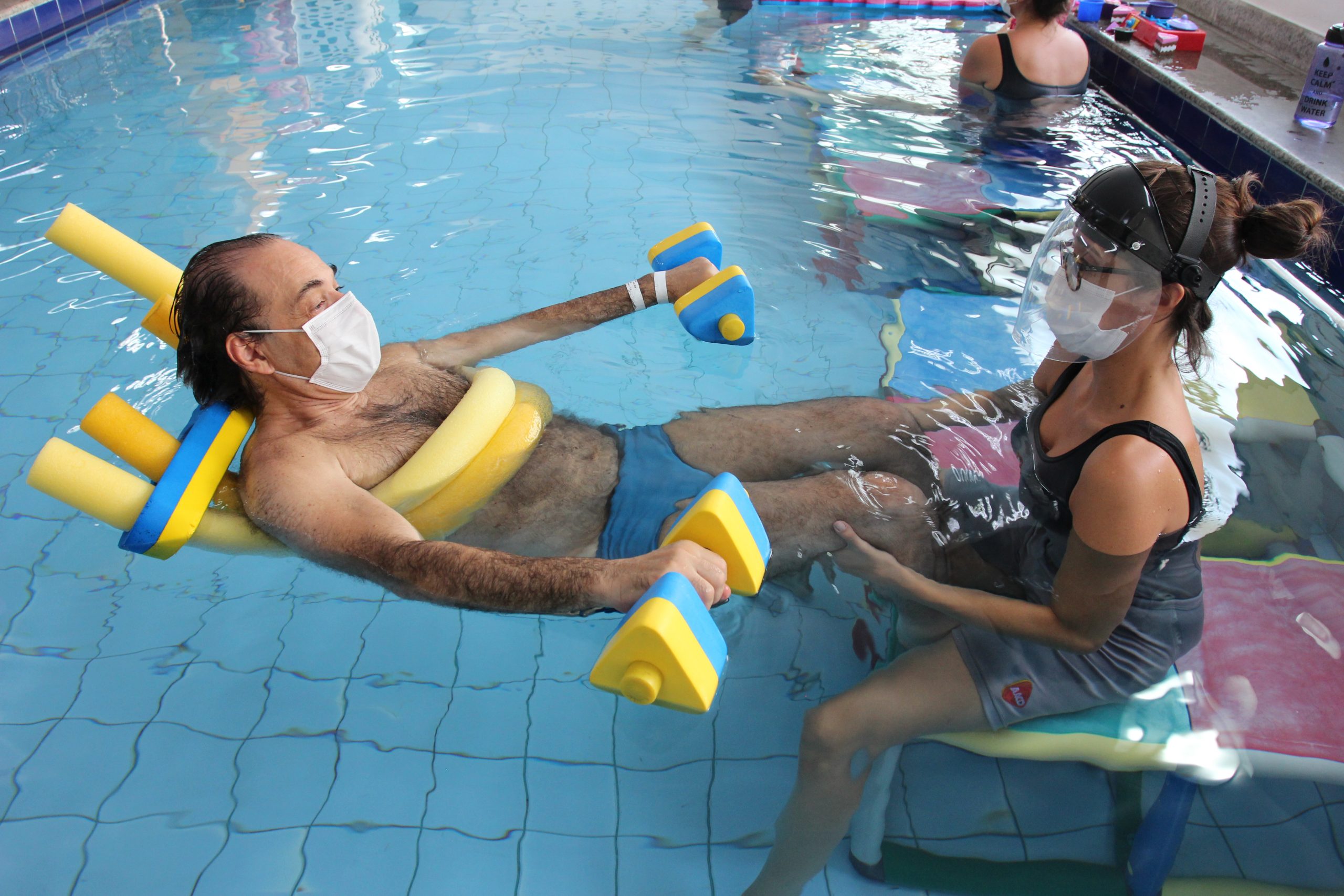 Homem com bóias em uma piscina, sendo auxiliado por uma profissional da Fisioterapia Aquática AACD