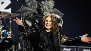 Ozzy Osbourne sentado em uma cadeira com design de morcego no último show do Black Sabbath. Ele é branco, tem cabelos compridos e está sorrindo de braços abertos.