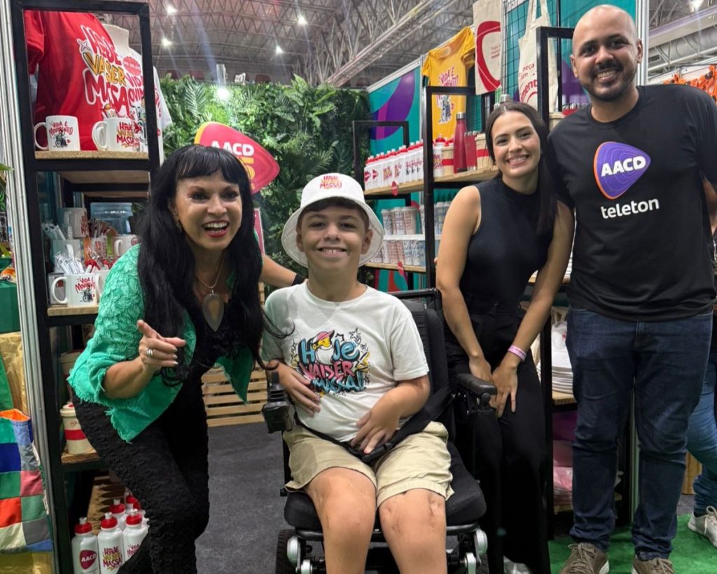 Quatro pessoas sorrindo na frente do estande da AACD Recife. Atrás vesse produtos estilizados pelo artista Véio Art na Fenearte. Na foto, há um menino na cadeira de rodas usando camiseta e chapéu bucket ilustrados pelo artista.