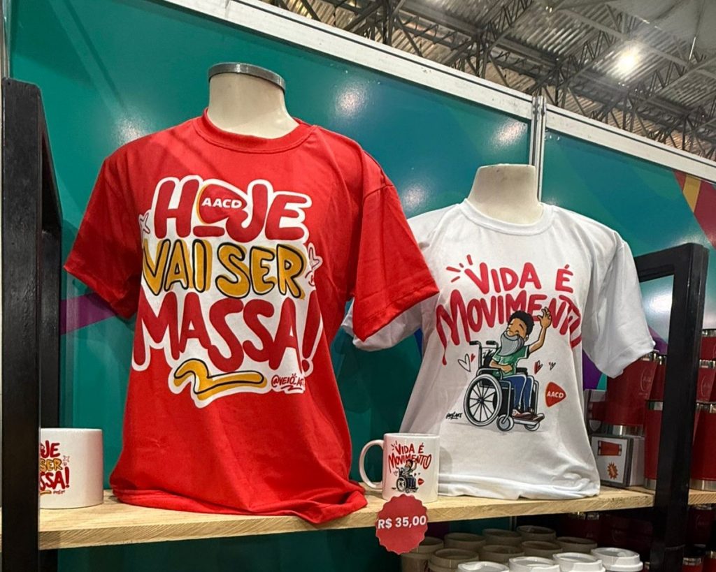 Foto de manequins com camisetas estampadas pelo artista Véio Art. Uma é vermelha com os dizeres "Hoje vai ser massa" e a outra é branca ilustrada com desenho de uma pessoa em cadeira de rodas e a frase "Vida É Movimento".