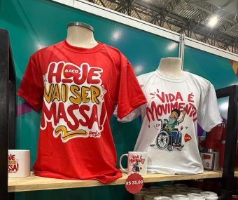 Foto de manequins com camisetas estampadas pelo artista Véio Art. Uma é vermelha com os dizeres "Hoje vai ser massa" e a outra é branca ilustrada com desenho de uma pessoa em cadeira de rodas e a frase "Vida É Movimento".