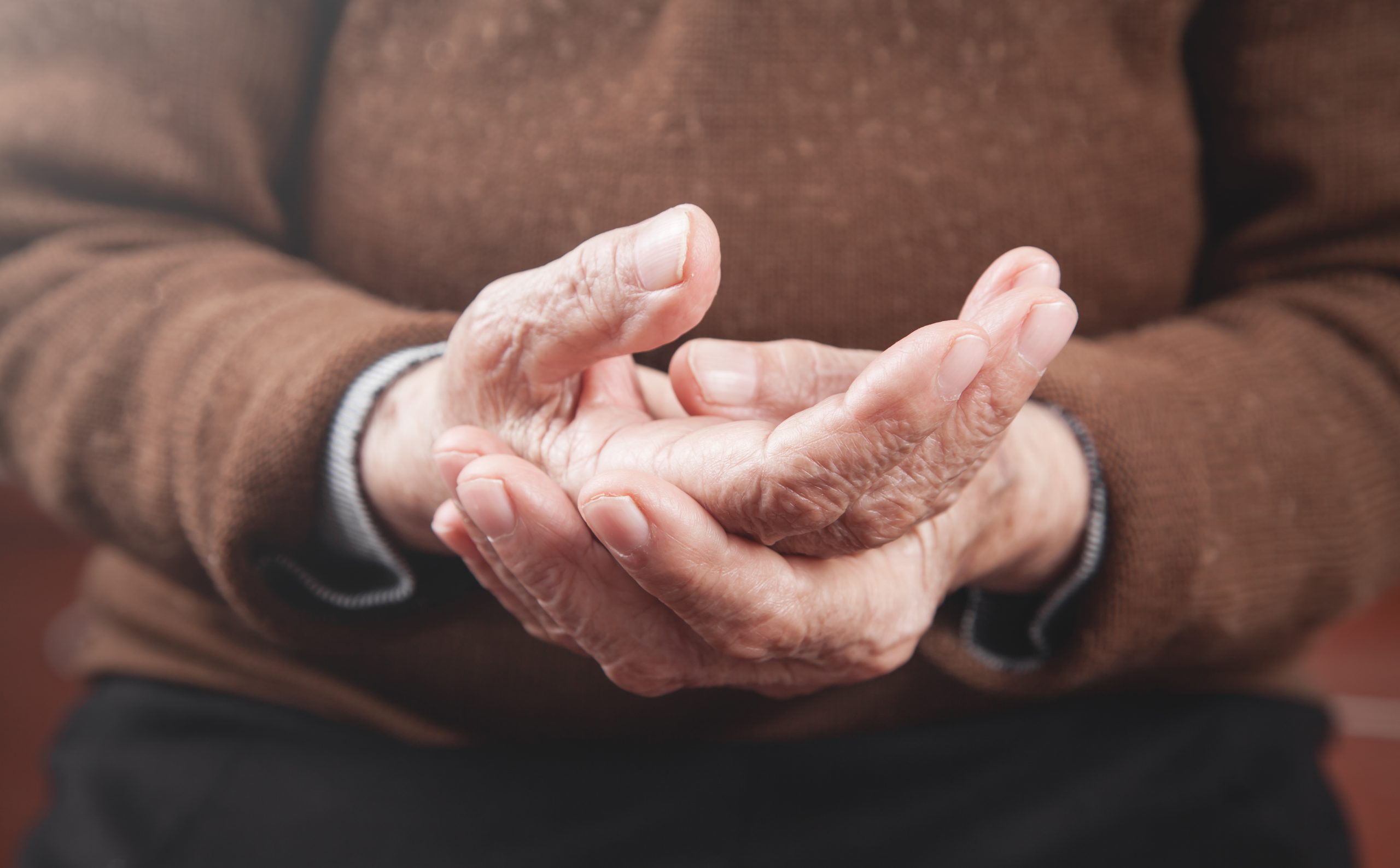 Pessoa idosa de pele branca segurando nas próprias mãos. Um dos sintomas do Parkinson.