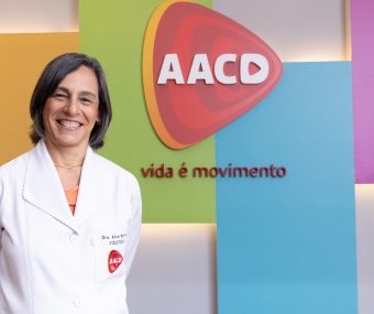 Dra. Alice Rosa Ramos, médica especializada em Fisiatria, de jaleco atrás de fundo colorido com logo da AACD