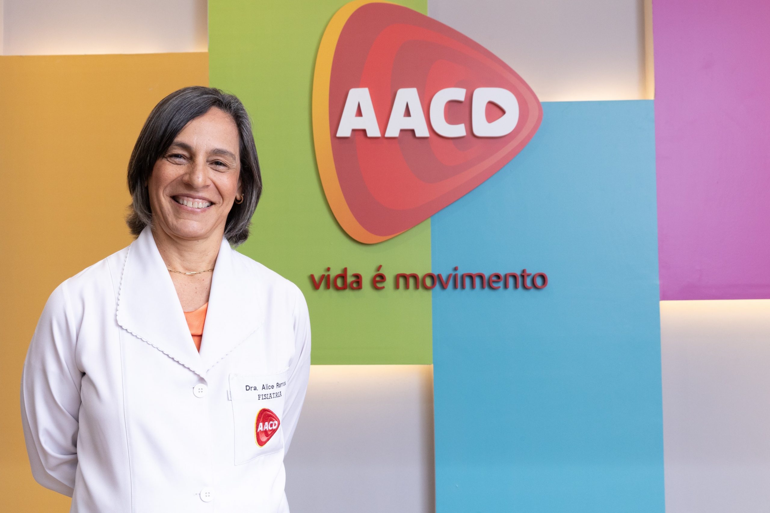 Dra. Alice Rosa Ramos, médica especializada em Fisiatria, atrás de fundo colorido com logo da AACD