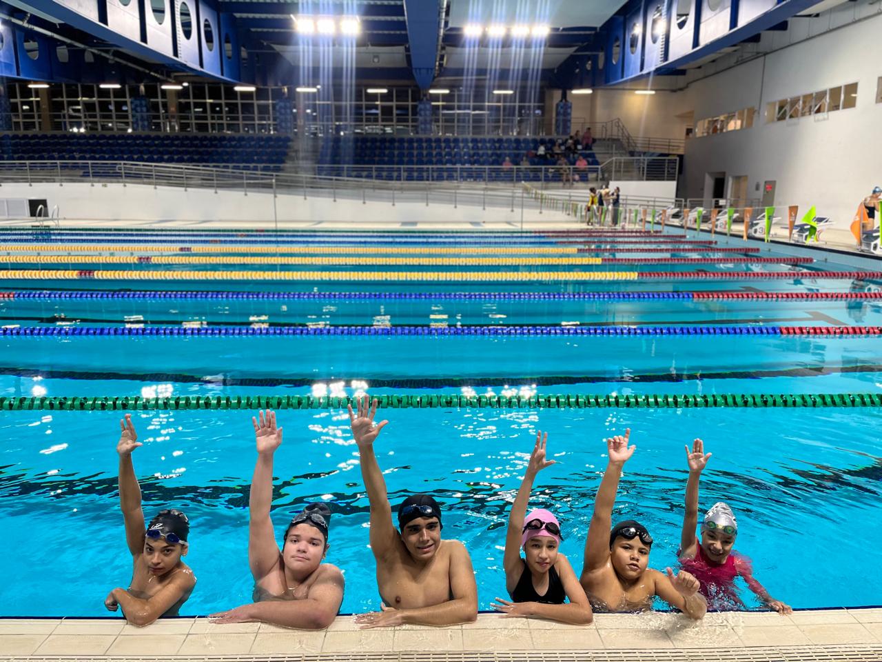 Atletas da AACD Esportes com os braços levantados na borda da piscina olimpica.