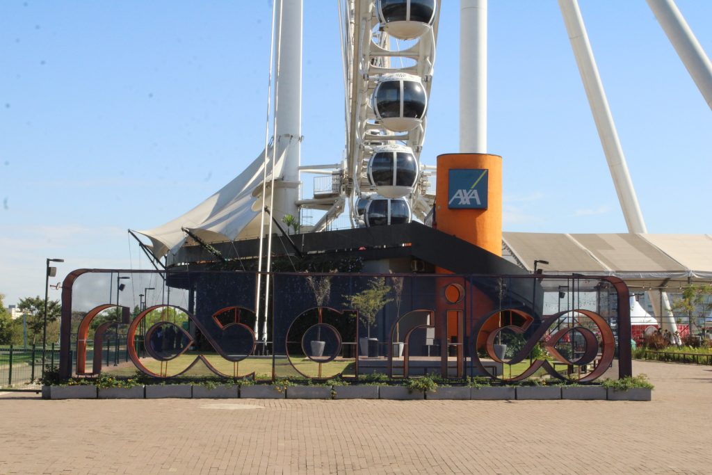 Foto de perfil da roda gigante do evento. Na frente um letreiro grande escrito Roda Rico