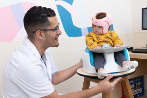Funcionário da Oficina Ortopédica da AACD ao lado de um bebê. A criança está sentada olhando para o adulto. Ela está usando uma órtese craniana feita sob medida para ela. 