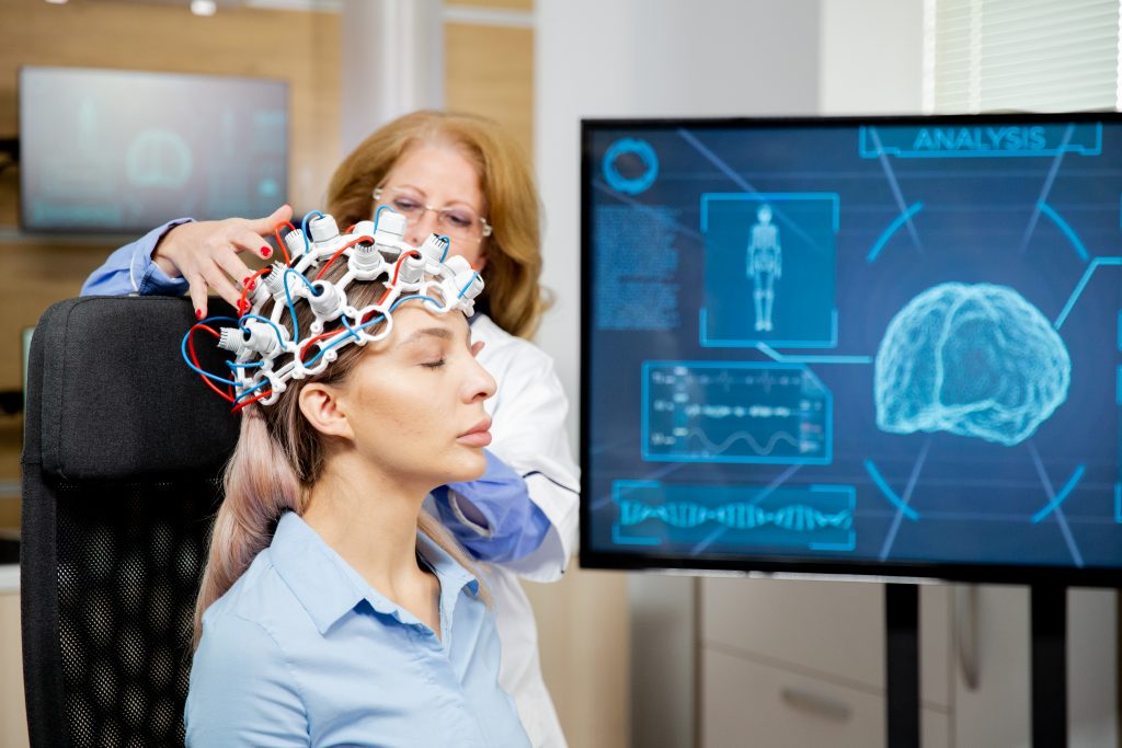 Profissional de saúde ajusta uma touca com eletrodos na cabeça de uma paciente sentada, durante um exame de atividade cerebral. Ao lado, um monitor exibe a representação de um cérebro e gráficos relacionados ao monitoramento neurológico.