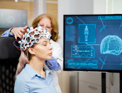 Profissional de saúde ajusta uma touca com eletrodos na cabeça de uma paciente sentada, durante um exame de atividade cerebral. Ao lado, um monitor exibe a representação de um cérebro e gráficos relacionados ao monitoramento neurológico.