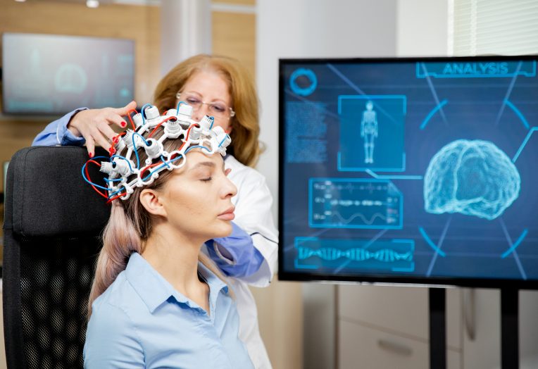 Profissional de saúde ajusta uma touca com eletrodos na cabeça de uma paciente sentada, durante um exame de atividade cerebral. Ao lado, um monitor exibe a representação de um cérebro e gráficos relacionados ao monitoramento neurológico.