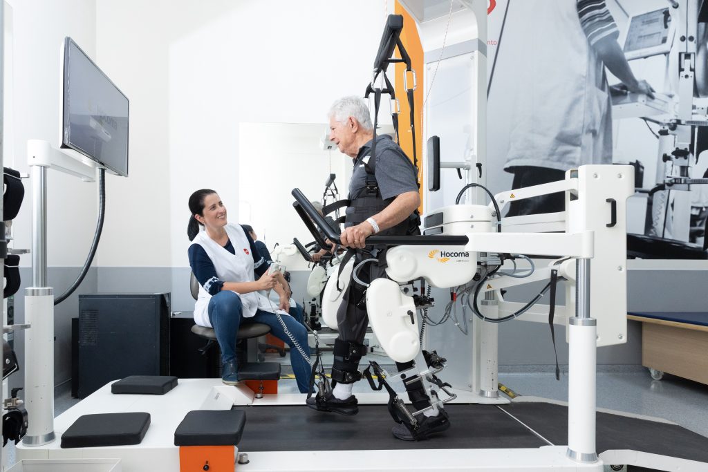 Homem idoso realiza treino de marcha em esteira com auxílio de equipamento robótico, enquanto profissional de saúde acompanha o exercício em sala de reabilitação.