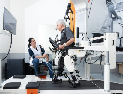 Homem idoso realiza treino de marcha em esteira com auxílio de equipamento robótico, enquanto profissional de saúde acompanha o exercício em sala de reabilitação.