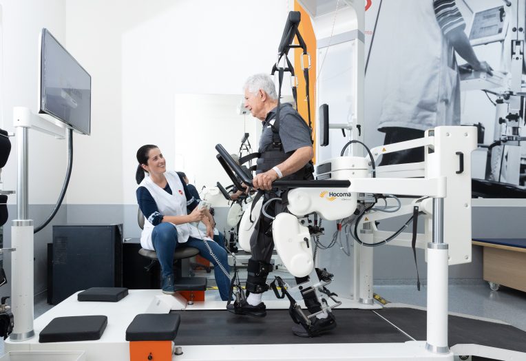 Homem idoso realiza treino de marcha em esteira com auxílio de equipamento robótico, enquanto profissional de saúde acompanha o exercício em sala de reabilitação.