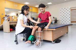 Profissional da AACD acompanha adolescente sentado em uma maca durante atividade de reabilitação, com dispositivo ortopédico na perna.